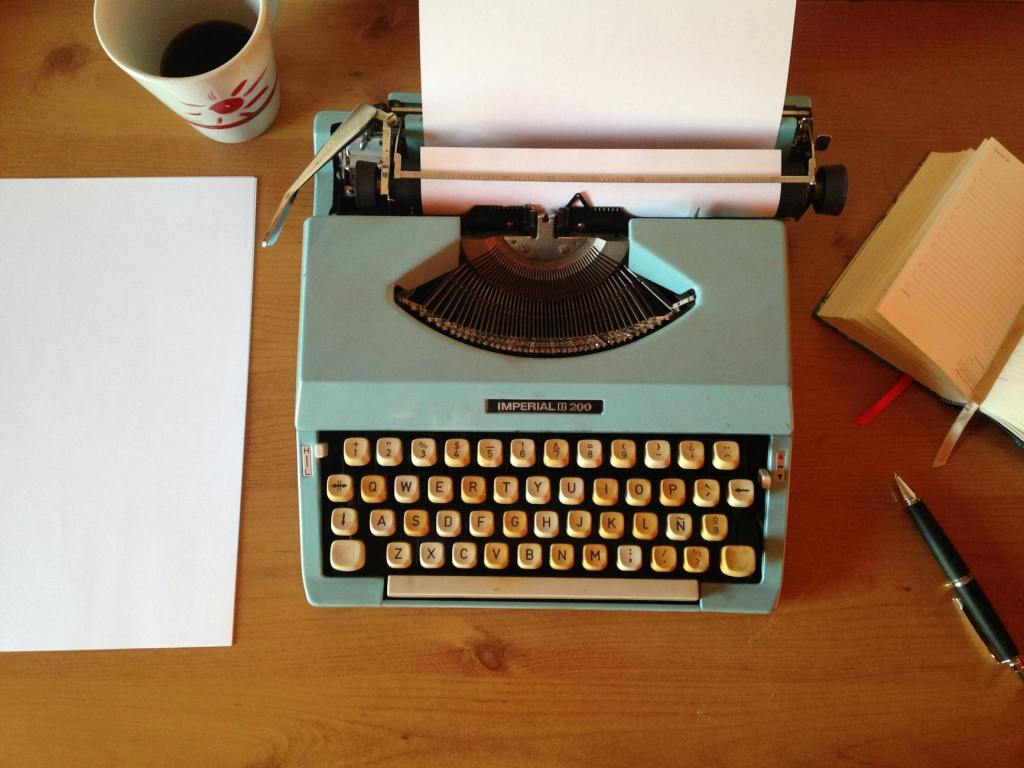 A brightly coloured retro typewriter is seen from above, with a blank piece of paper inserted and one to the side. A cup is just visible.