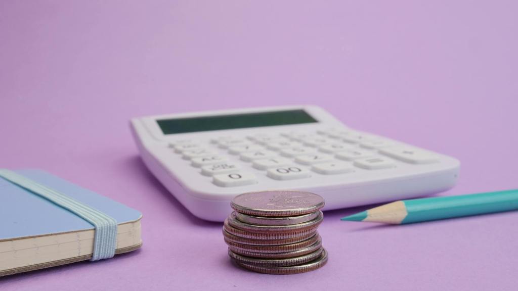 On a lilac coloured background a calculator is slightly out of focus with a pile on coins in front, a sharp blue pencil and a notebook slightly obscured.