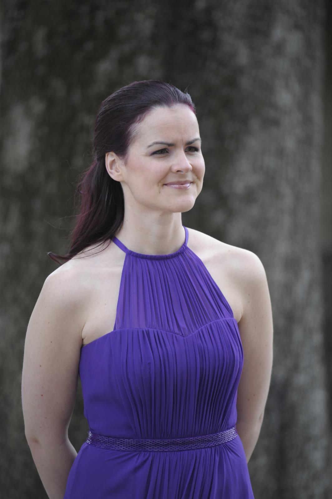A younger image of Dawn from hips up in front of a blurred back ground.  Dawn wears a roman style deep purple dress, with smooth hair sleeked back from her face and flowing down her back. 
