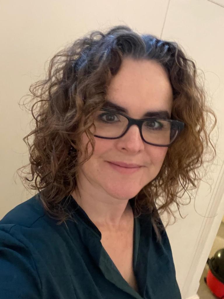 A headshot or selfie of a lady in her early forties. She has brown hair in messy curls to her shoulders, and pale skin, she wears glasses and a green blouse, casually open at the next. In the lower corner of the image a vacuum cleaner can be glimpsed in the background.