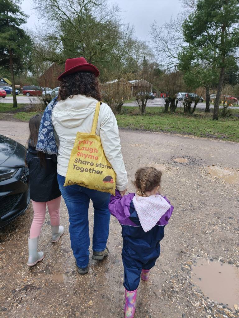 View from behind of Dawn walking holding hands with two girls.  They walk across a muddy/potholed carpark. towards distant trees and buildings.