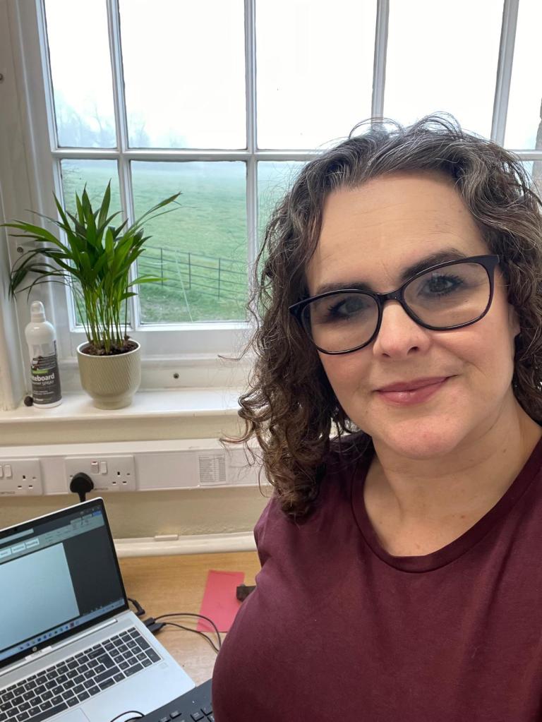 Image of Dawn, a lady in her early 40s, shown from chest up.  Her dark and grey scattered hair sits in messy curls to her shoulders and she wears glasses and a simple burgundy coloured t-shirt.
Behind her is a large sash window looking over a field.  A plant sits on the windowsill over a desk where Dawn's laptop sits with a blank document on the screen.